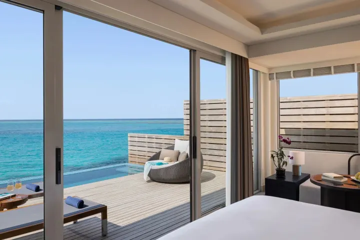 Avani+ Fares Maldives Resort — светлая спальня водной виллы с панорамным видом на лагуну