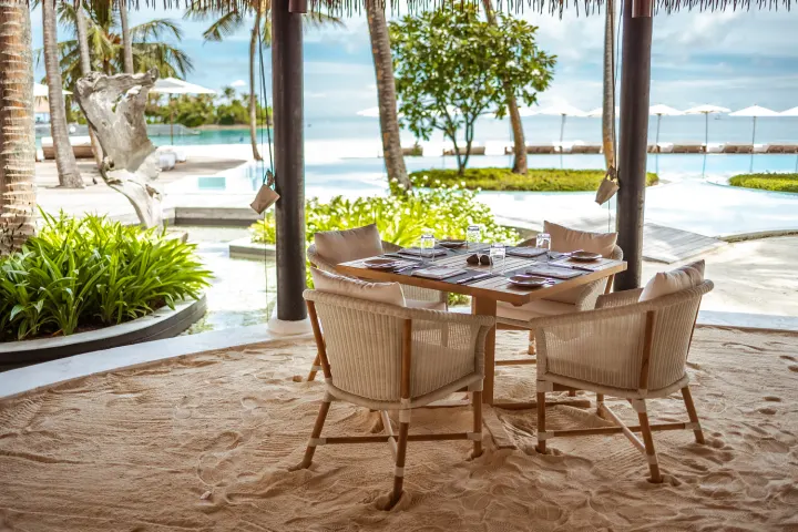 Maalifushi by COMO — столик в ресторане с песчаным полом и плетеными креслами