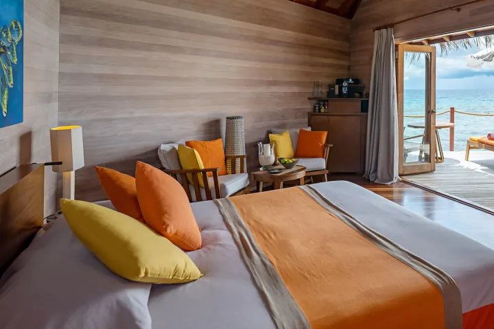 V Villas Maldives at Mirihi — светлая спальня водной виллы с отделкой из натурального дерева и видом на океан